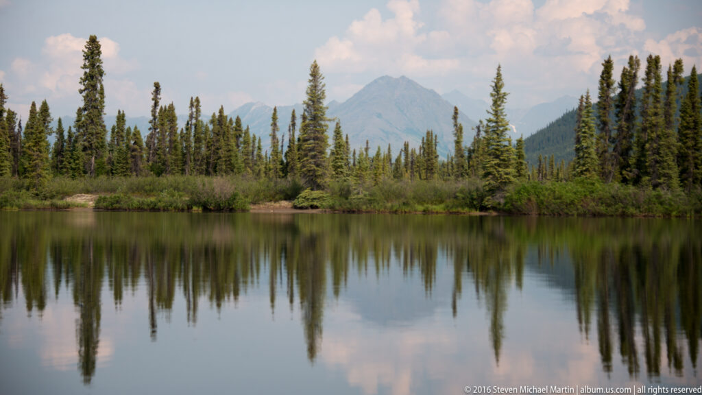 A pointed mountain peak rises behind spruce trees and is reflected with the shoreline in a calm lake.