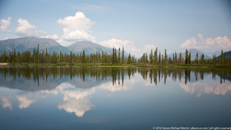 A wide lake mirrors a spruce shoreline, mountain range, and scattered clouds beneath a blue sky.
