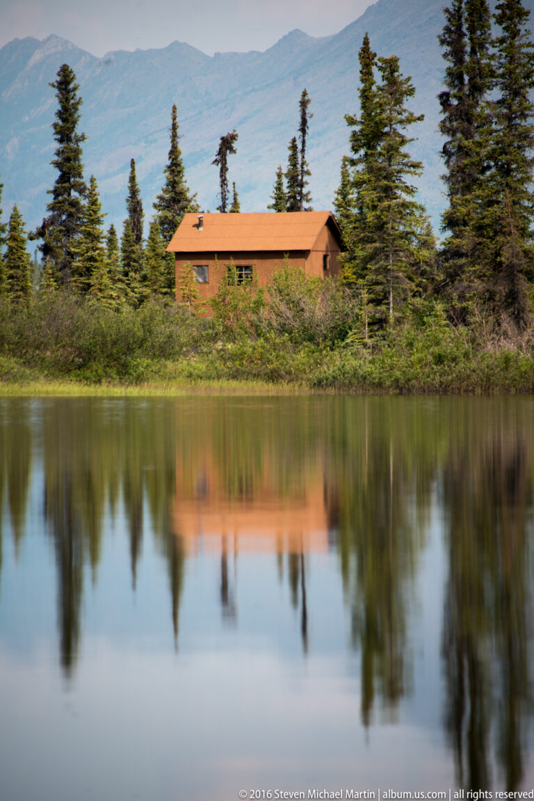 A small wooden cabin is partly screened by spruce and reflected in a still lake.