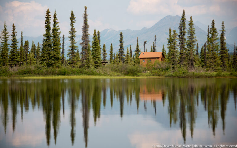 A small orange-brown cabin sits among spruce on the far shore, with its reflection visible in the lake.