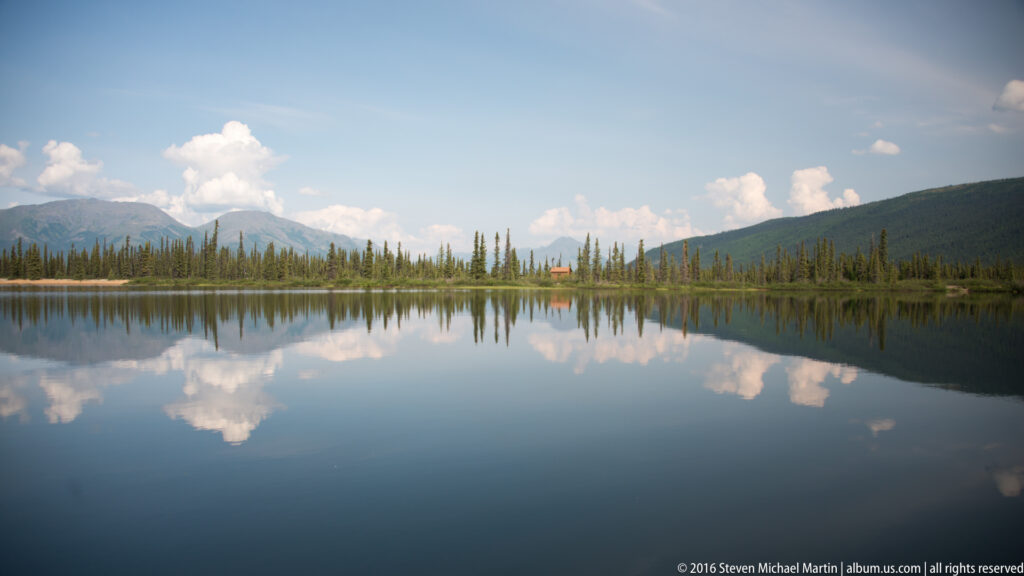 A wide lake, distant cabin, spruce shoreline, and mountain range sit beneath large clouds and reflect in the water.