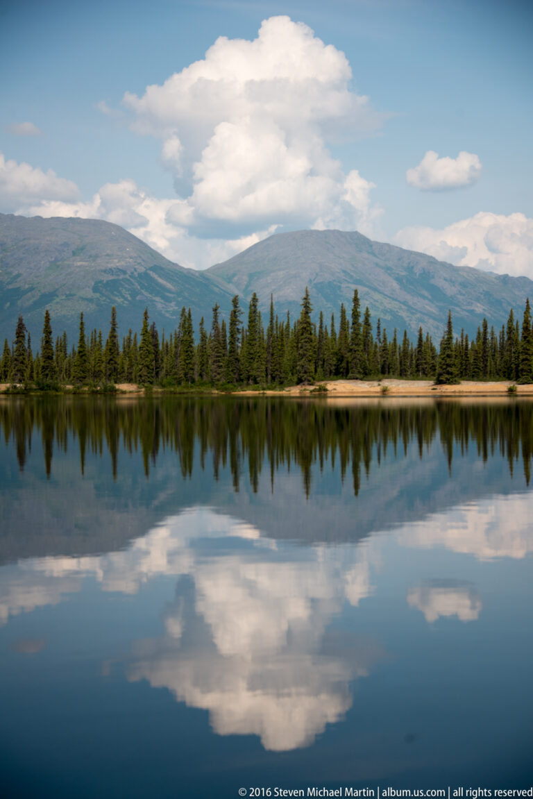 A broad lake reflects spruce forest, mountain ridges, and large white clouds beneath a blue sky.