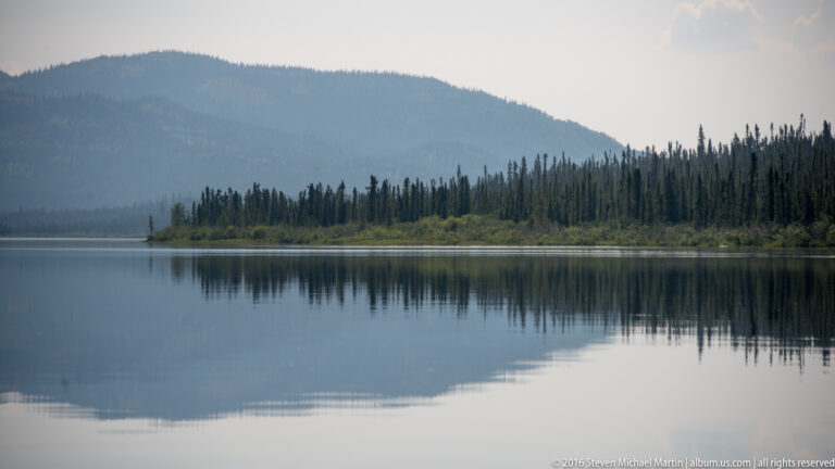 A spruce-covered shore and hazy mountain slope form a soft reflection in a calm lake.