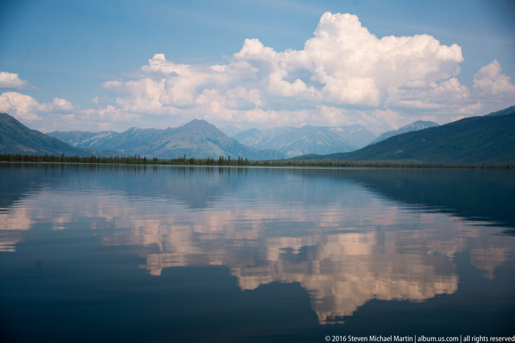 A tall cumulus cloud and distant mountains are reflected in a broad lake framed by dark slopes.