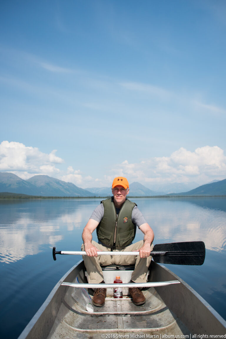 A man wearing an orange cap and flotation vest sits in a canoe, holding a paddle across his lap on a lake.