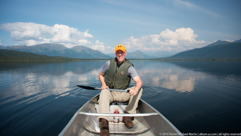 A man wearing an orange cap and life jacket sits in a canoe on a calm mountain lake.