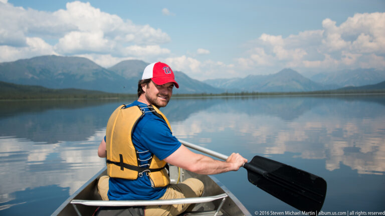 A man in a yellow life jacket paddles a canoe across a reflective lake.