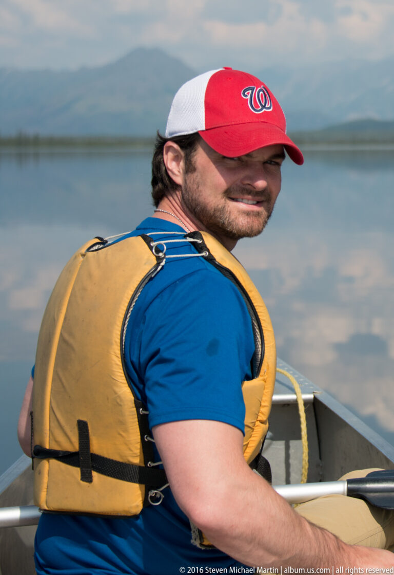 Close view of a man in a yellow life jacket and red-and-white cap turning toward the camera in a canoe.