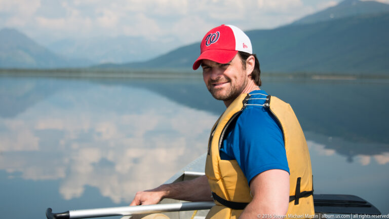 A man in a yellow life jacket sits in a canoe with a paddle, looking toward the camera.