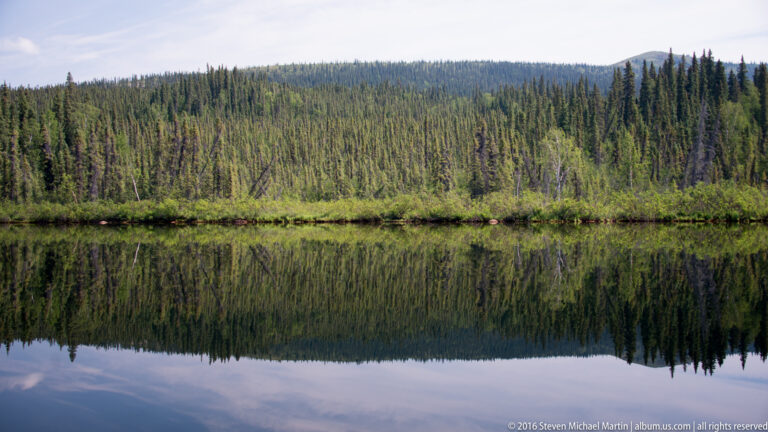 A forested shoreline and mountain slope reflected in still lake water.