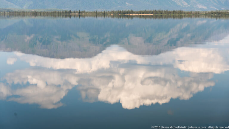 Bright clouds and distant mountain silhouettes reflected in a calm blue lake.