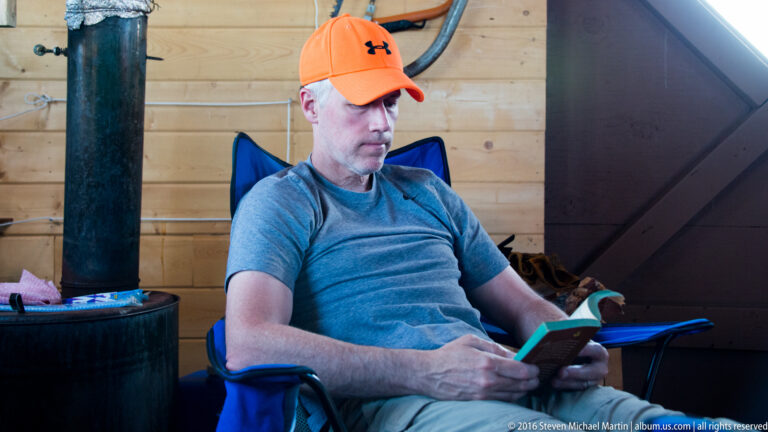 A man in an orange cap sits reading beside a black wood stove in a wood-paneled cabin.