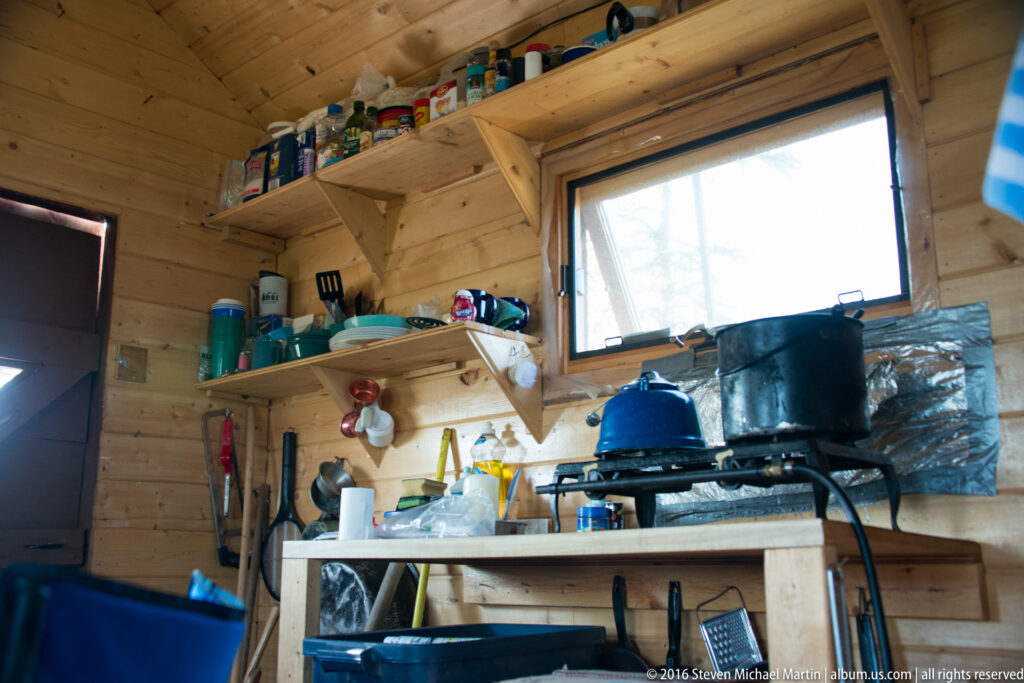 Blue kettle and black pot sit on a freestanding stove beneath shelves in a small wood cabin kitchen.