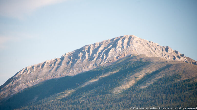 Sunlit rocky mountain summit above a dark forested slope under a blue sky.
