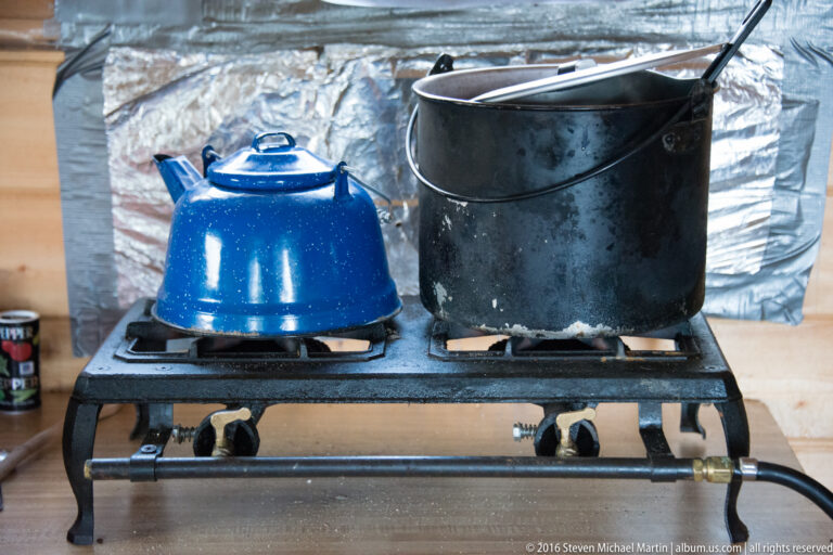 Blue enamel kettle and black pot on a two-burner camp range inside a sheltered space.