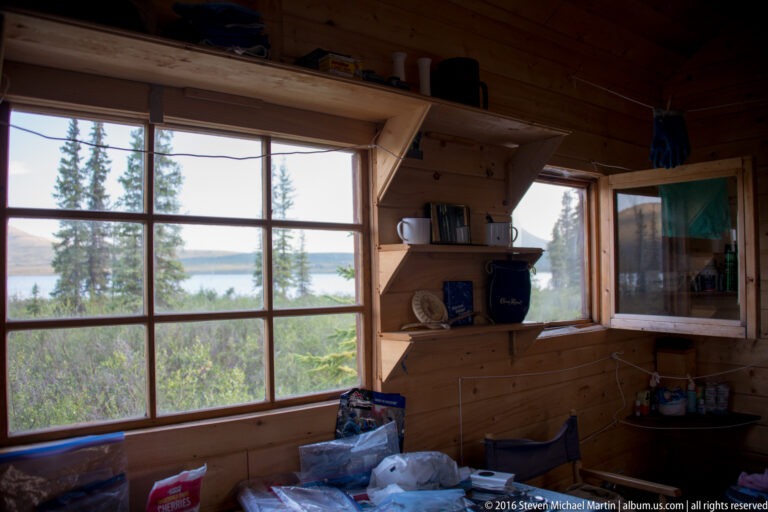 Wood cabin interior with large windows overlooking trees and a lake, with supplies on a counter.