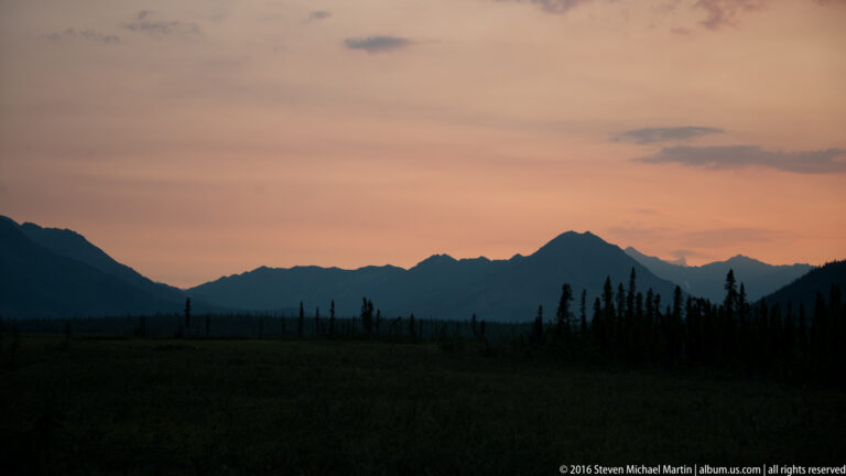 Pink-orange sunset above dark mountain ridges and a line of spruce trees.