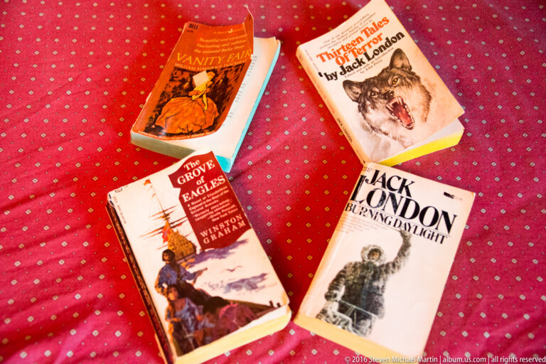 Four paperback adventure books, including two by Jack London, lie on a red patterned bedspread.
