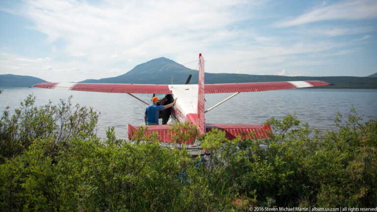 Red-and-white floatplane at the lake’s edge as a person stands beside its open cabin.