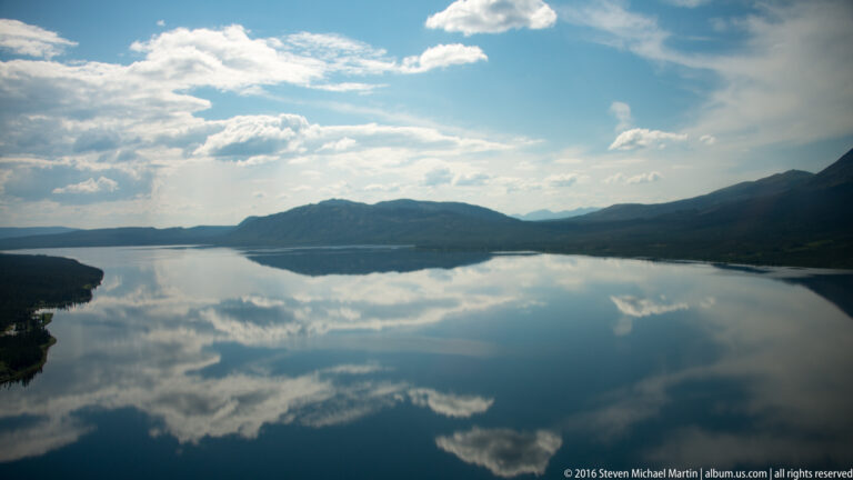 Clouds and low mountain ridges reflect across a broad lake beneath a blue sky.