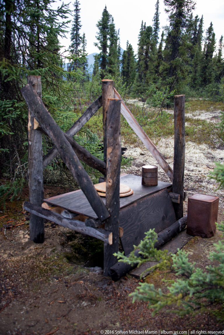 A weathered timber framework with crossed beams and wooden boxes stands in a sandy clearing beside spruce forest.