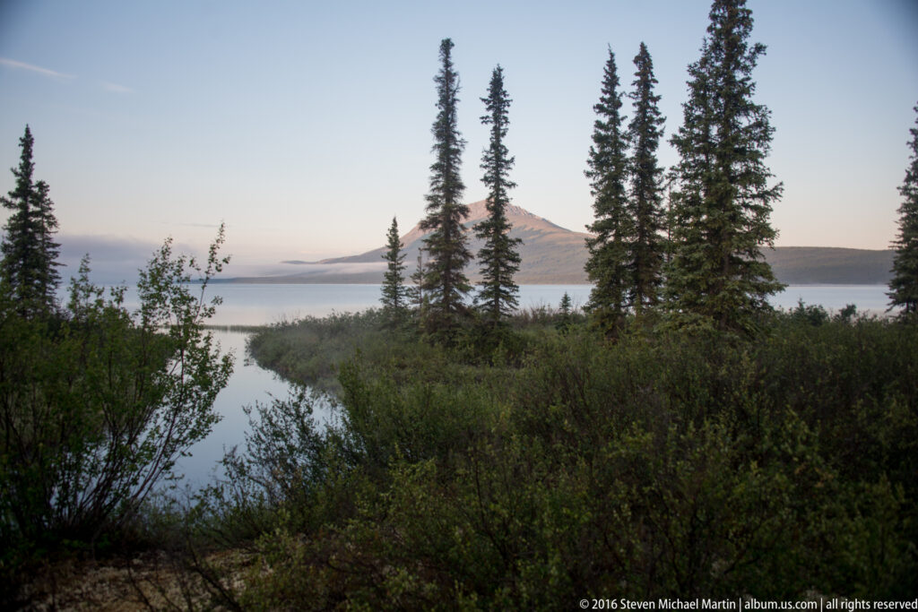 A narrow channel through lakeside shrubs opens onto a lake and a distant mountain in warm light.