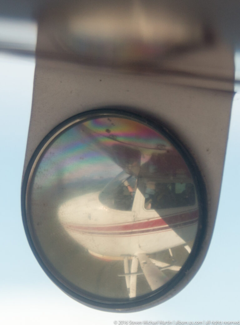 A red-and-white floatplane is reflected in an oval glass surface against a pale blue background.