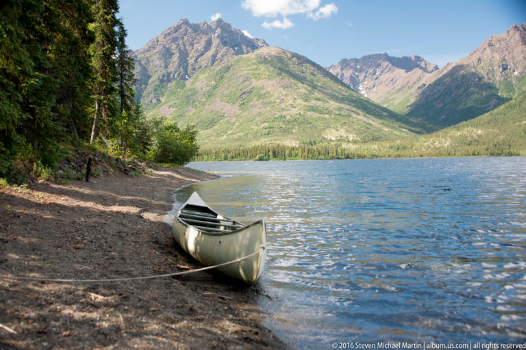 A green canoe is pulled onto a pebbled shore and tied with a rope beside a mountain lake.