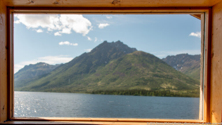A wooden cabin window frames a dark mountain and a strip of lake beneath a partly cloudy sky.
