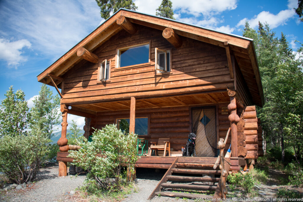 A two-story log cabin with a covered porch and steps stands among shrubs and spruce trees.