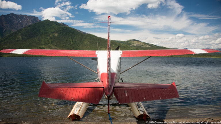 The rear of a red-and-white floatplane rests on the lake, its wings spanning the mountain-lined water.