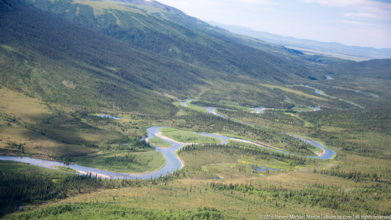 An aerial view shows a winding river crossing a broad green valley between forested mountain slopes.