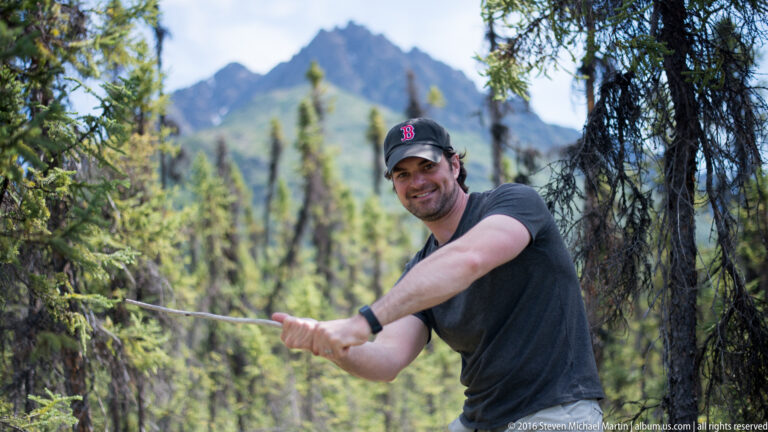 A smiling man in a dark shirt and cap holds a long branch among trees, with a mountain behind him.