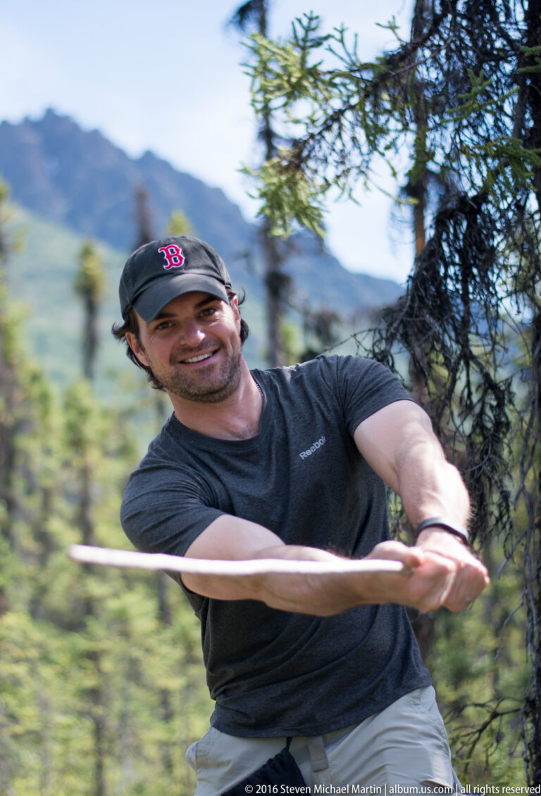 A smiling man in a dark cap holds a thin branch across the foreground against blurred green trees.