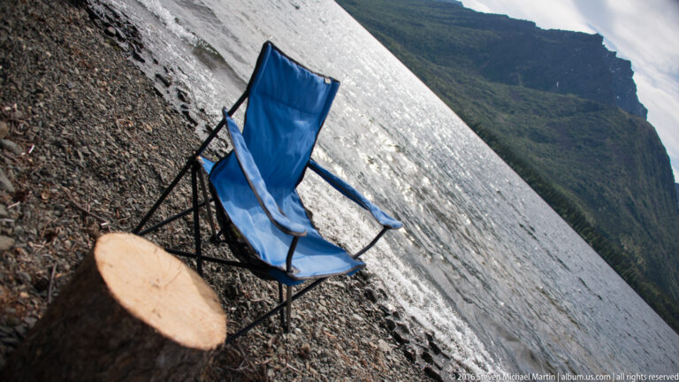 A blue folding chair and cut log sit on a steep rocky slope above a green mountain valley.