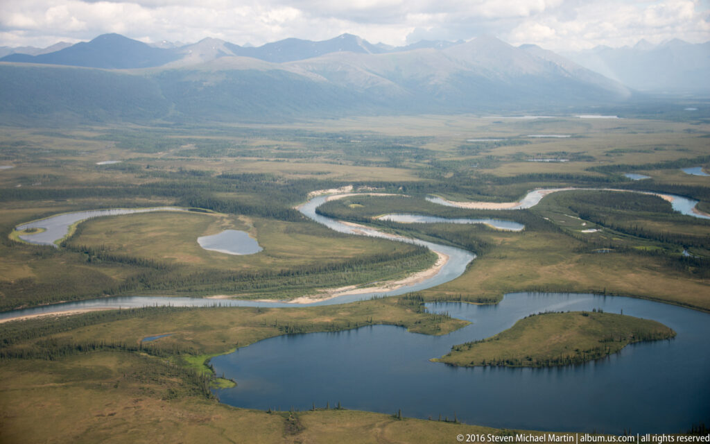 A broad river curves through open tundra past small ponds and into a larger lake, with mountains in the distance.