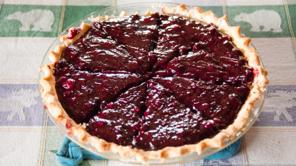 A round berry pie with a crimped crust and glossy dark fruit rests on a patterned cloth.
