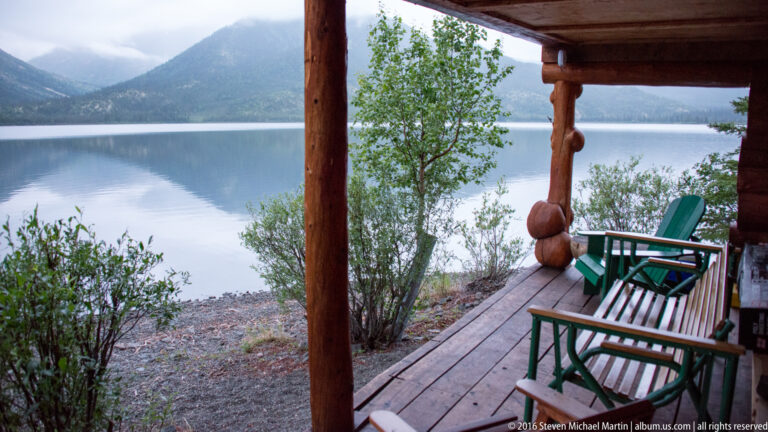 Green chairs on a log cabin porch face a calm lake and distant wooded hills.