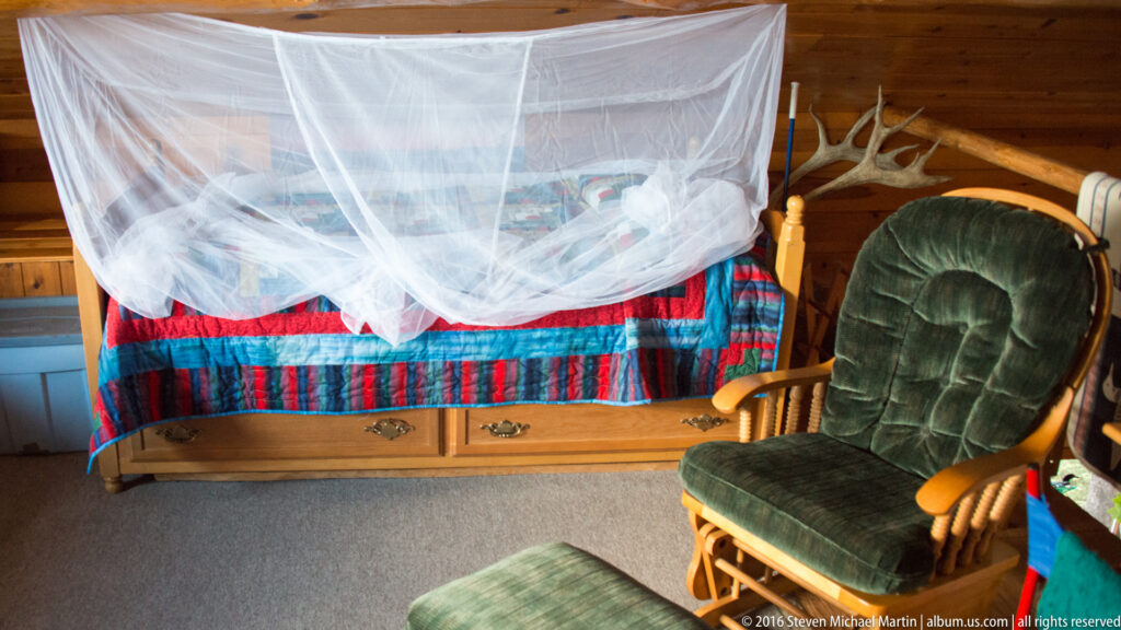 White mosquito net drapes over a colorful bed beside a green rocking chair in a wood cabin.