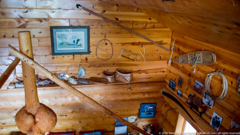 Wood cabin wall with a framed picture, coiled rope, snowshoes, and assorted equipment.