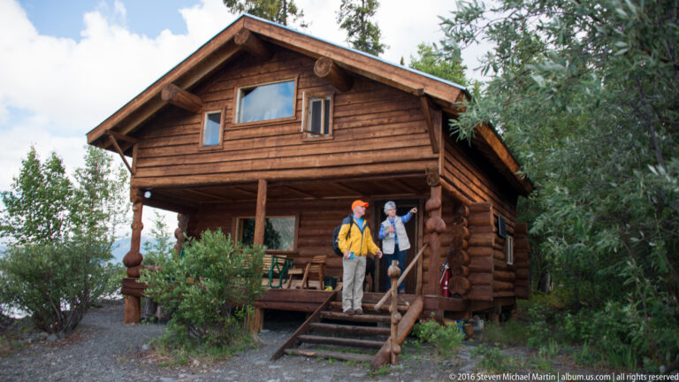 Two visitors stand on the steps of a two-story log cabin beside green porch chairs.