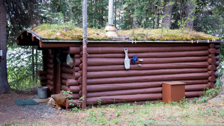 Low log cabin with a moss-covered roof, stovepipe, and objects hanging on its wall.