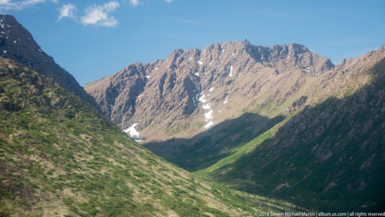 A steep rocky peak rises above green valley slopes, with small snow patches remaining in its gullies.