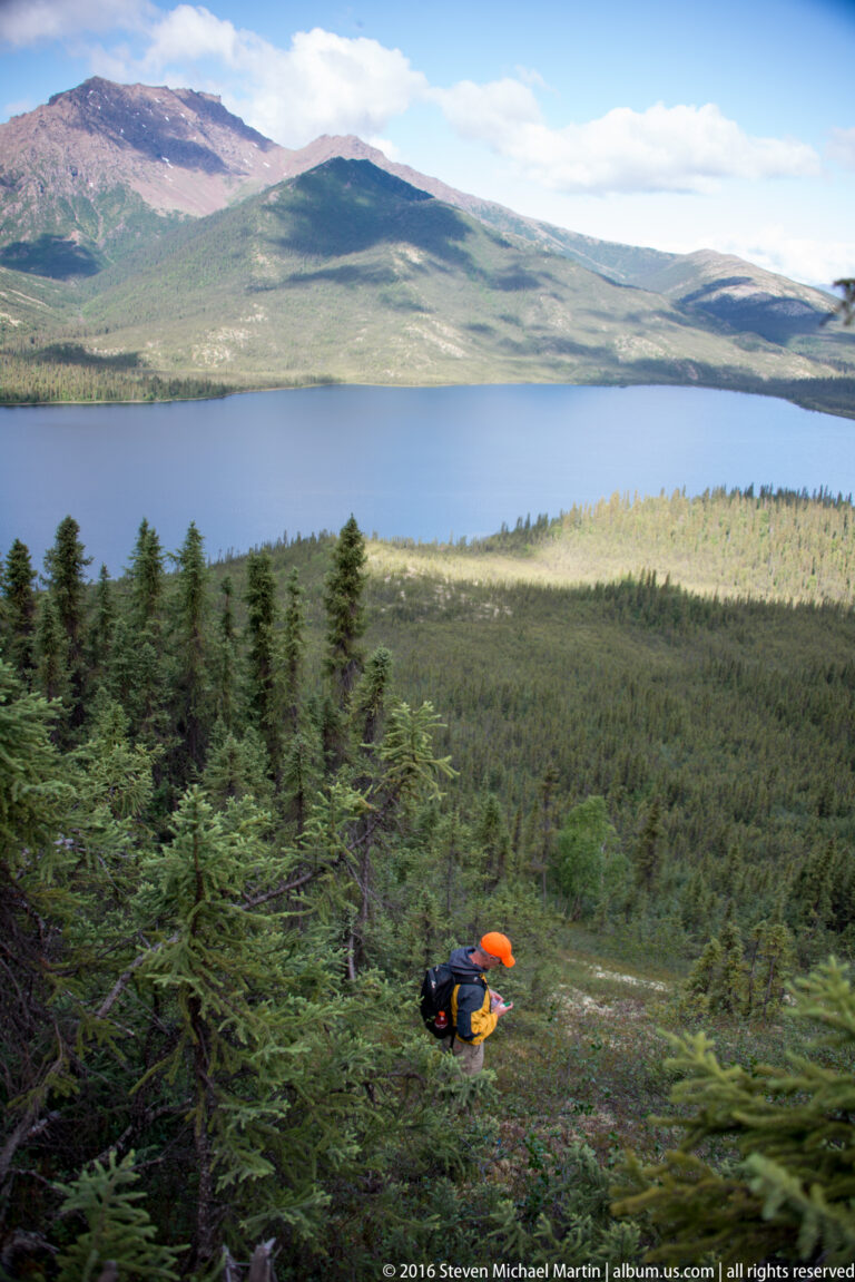 A small hiker stands among brush above a broad lake and rolling hills.