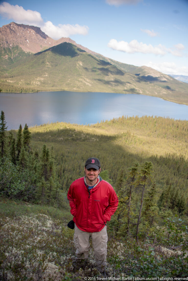 A man in a red jacket stands on a brushy slope above a lake and distant hills.