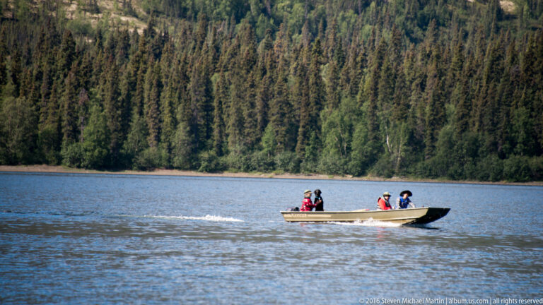 Three people ride in a small motorboat across a lake, leaving a wake beside a forested shore.