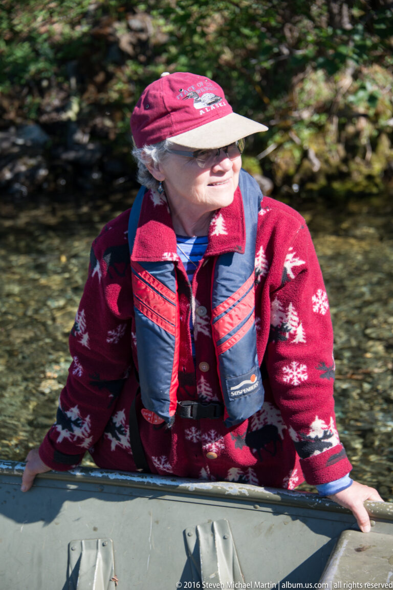 Woman in a red patterned sweater and blue life vest seated in a canoe on clear water.