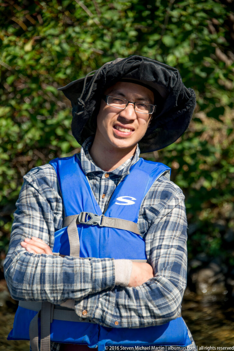 Smiling man in glasses, plaid shirt, blue life vest, and wide-brim hat before green foliage.