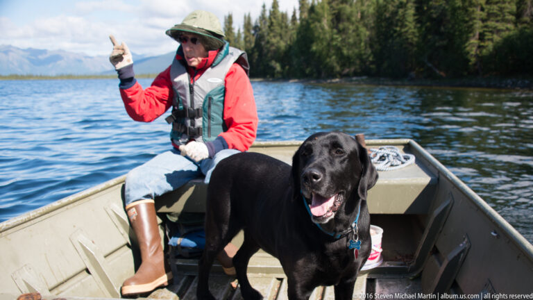 Woman in a life vest sits in a canoe with a black dog on the lake.