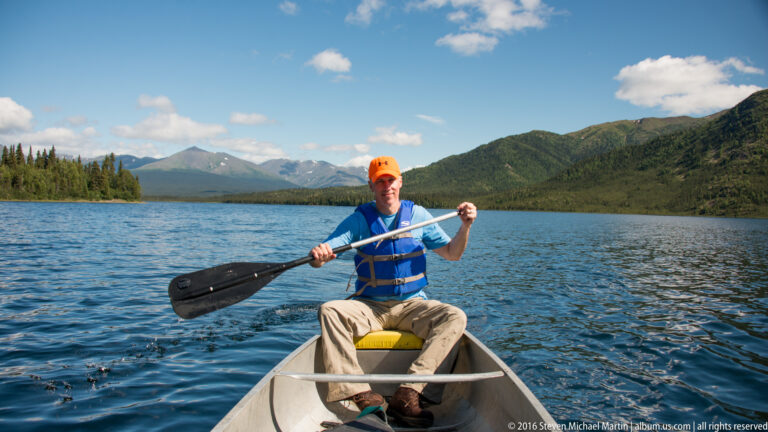 Man in an orange cap and blue life vest paddles an aluminum canoe on a mountain lake.